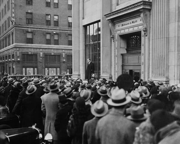 A black-and-white photograph of a crowd, mostly made up of men in hats, gathering outside a large building and listening to a speaker, who is holding his hat in his hand and standing in front the building’s large ground floor window. The words “Dedicated to Thrift" appear above the building’s entrance.