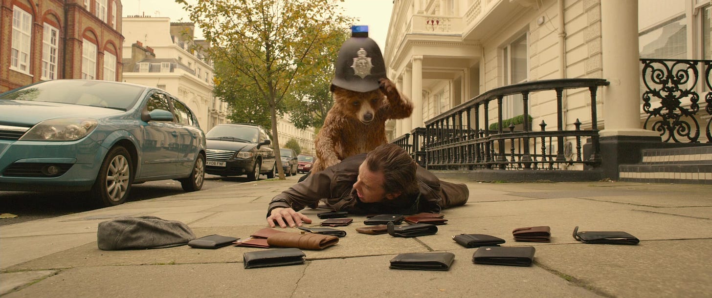 A still image from the first Paddington movie, in which Paddington Bear has just landed atop a pickpocket, spilling 16 wallets to the ground. Paddington is wearing a police helmet.