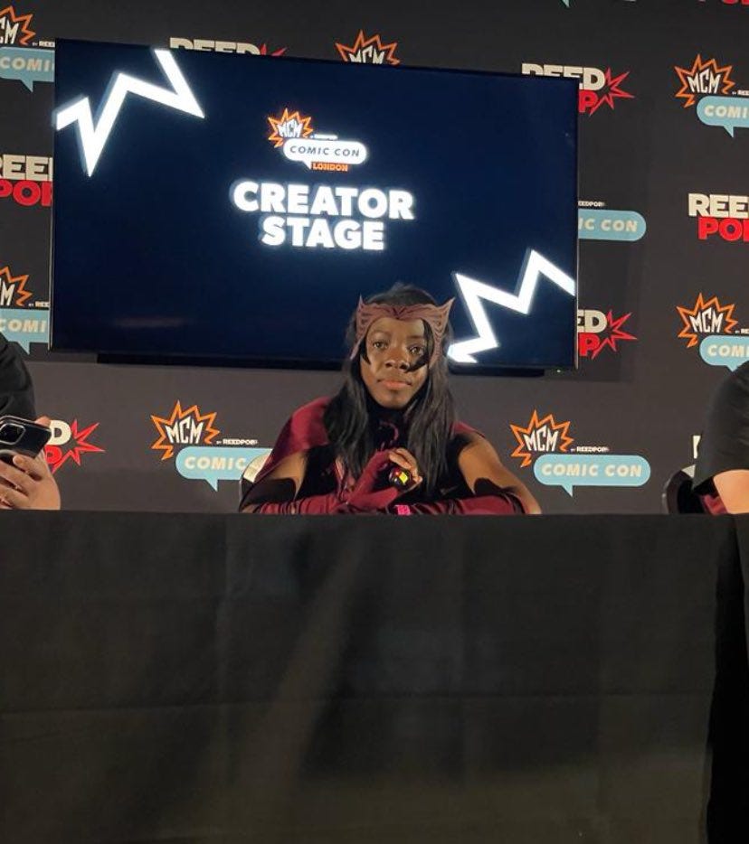 Myself dressed in Wanda cosplay, burgundy cape, and gloves and her witchy headband. Sat behind a desk infront of a screen that says 'creator stage' at Comic Con