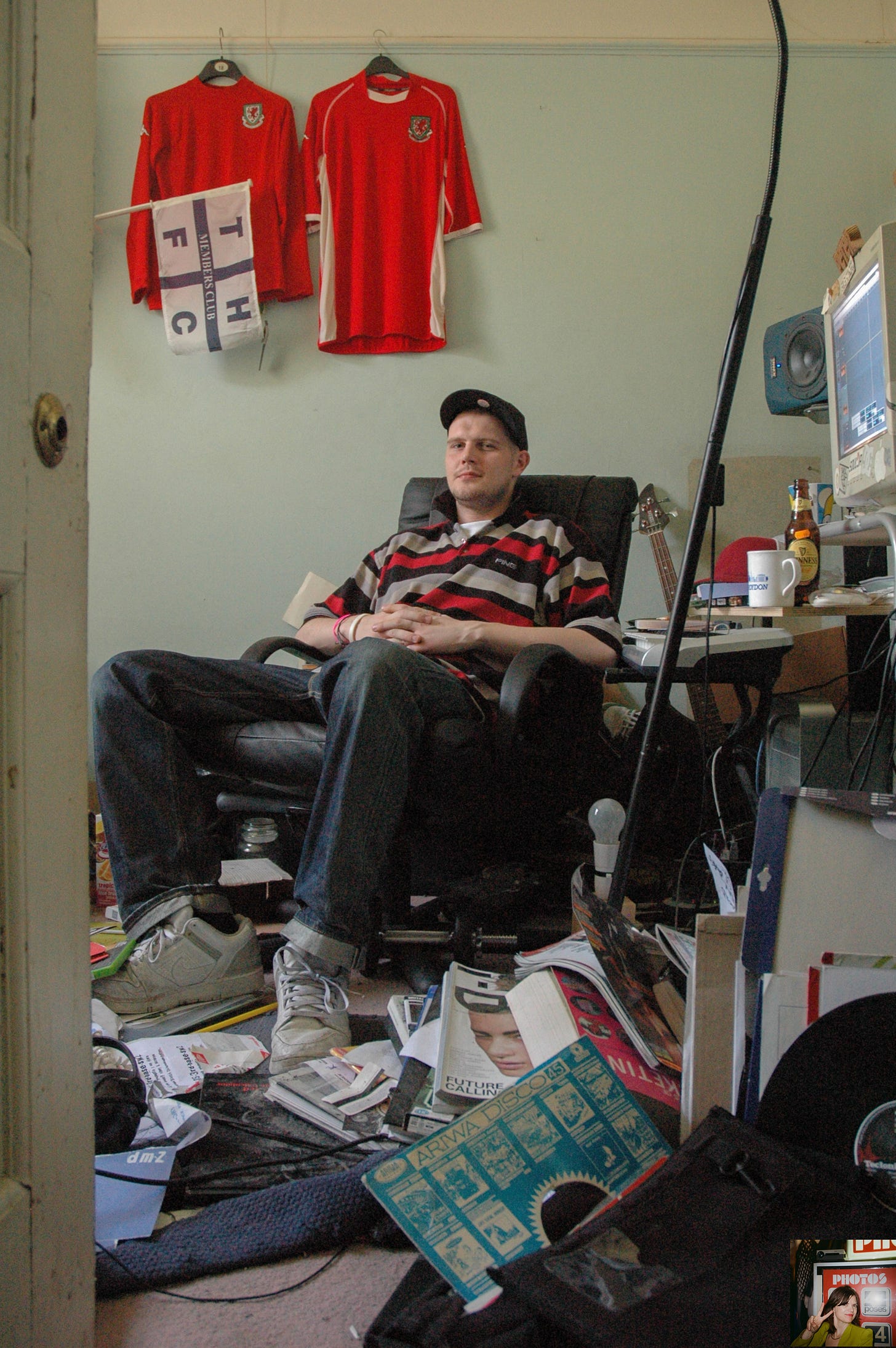 A young man called Loefah sat on an office chair looking directly at the camera in a room full of rugby shirts, a computer, drinks, instruments, records and other objects