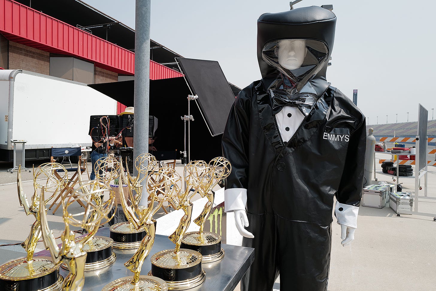 A mannequin in a hazmat suit that says "Emmy's" stands over some trophies. 
