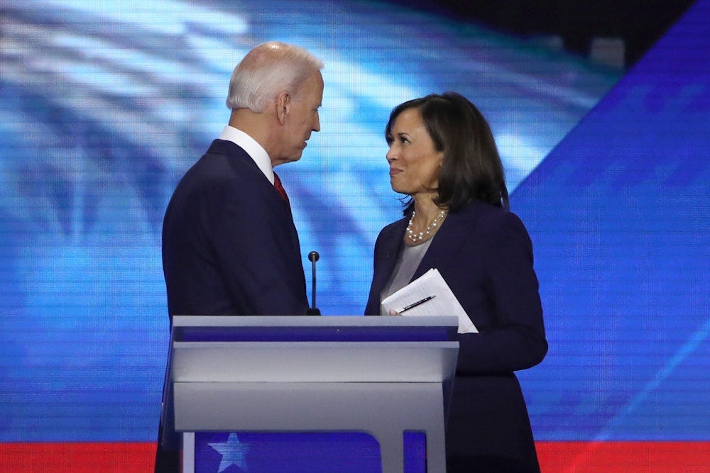 Kamala Harris looks up at Joe Biden and smiles as they stand behind a lectern in 2019
