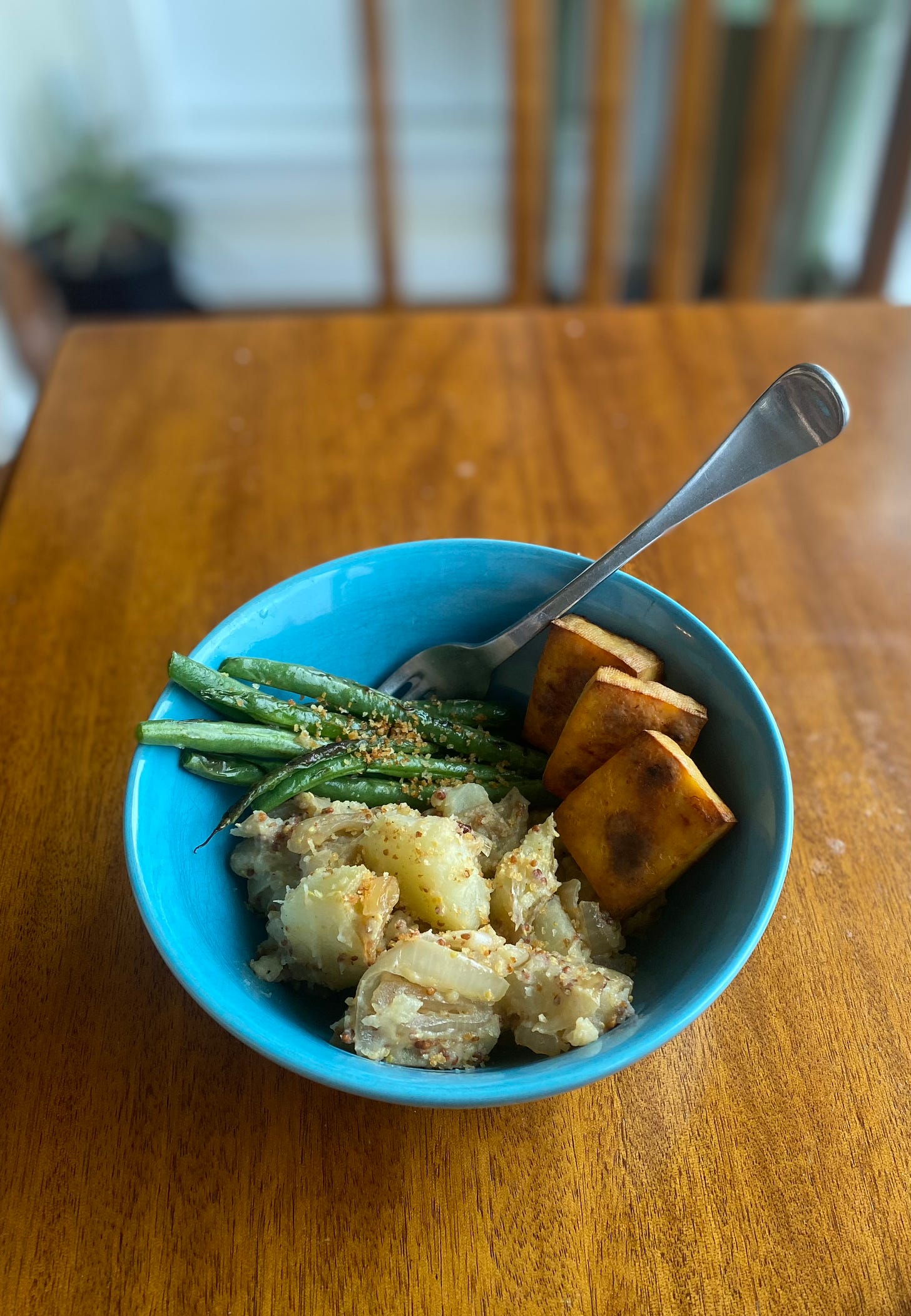 A blue bowl of the potato salad described above, with three squares of roasted tofu on one side, and seared green beans at the back. Toasted panko crumbs are sprinkled over the beans.