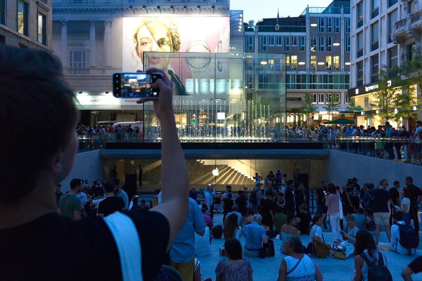 The amphitheater at Apple Piazza Liberty is full of activity shortly after the store opening. One person is taking a photo of the crowd with their iPhone.