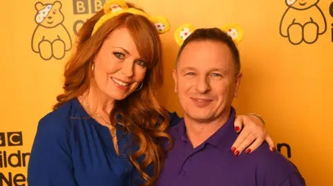 Getty 
Gabrielle Blackman (L) and Mark Millar of DIY SOS backstage at BBC Children in Need's 2019 Appeal night at Elstree Studios. Mark is wearing a purple shirt and Gabrille is wearing a navy dress, they both have Pudsey's ear headband on and are staying in front of Pudsey bear and BBC Children in Need logo on a yellow wall.
