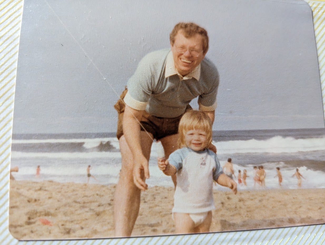 A father stands protectively over his child at the beach, both of them smiling, sand, waves, and other people in the background