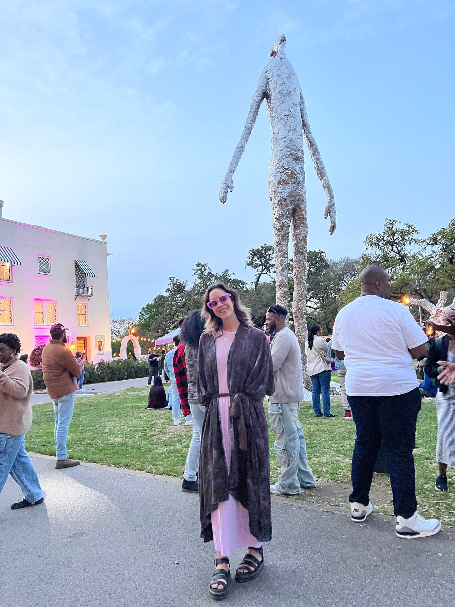 A white woman in a purple dress poses and smiles in front of a 20-foot-tall sculpture that looks like a silvery skinny alien man in front of a blue sky.