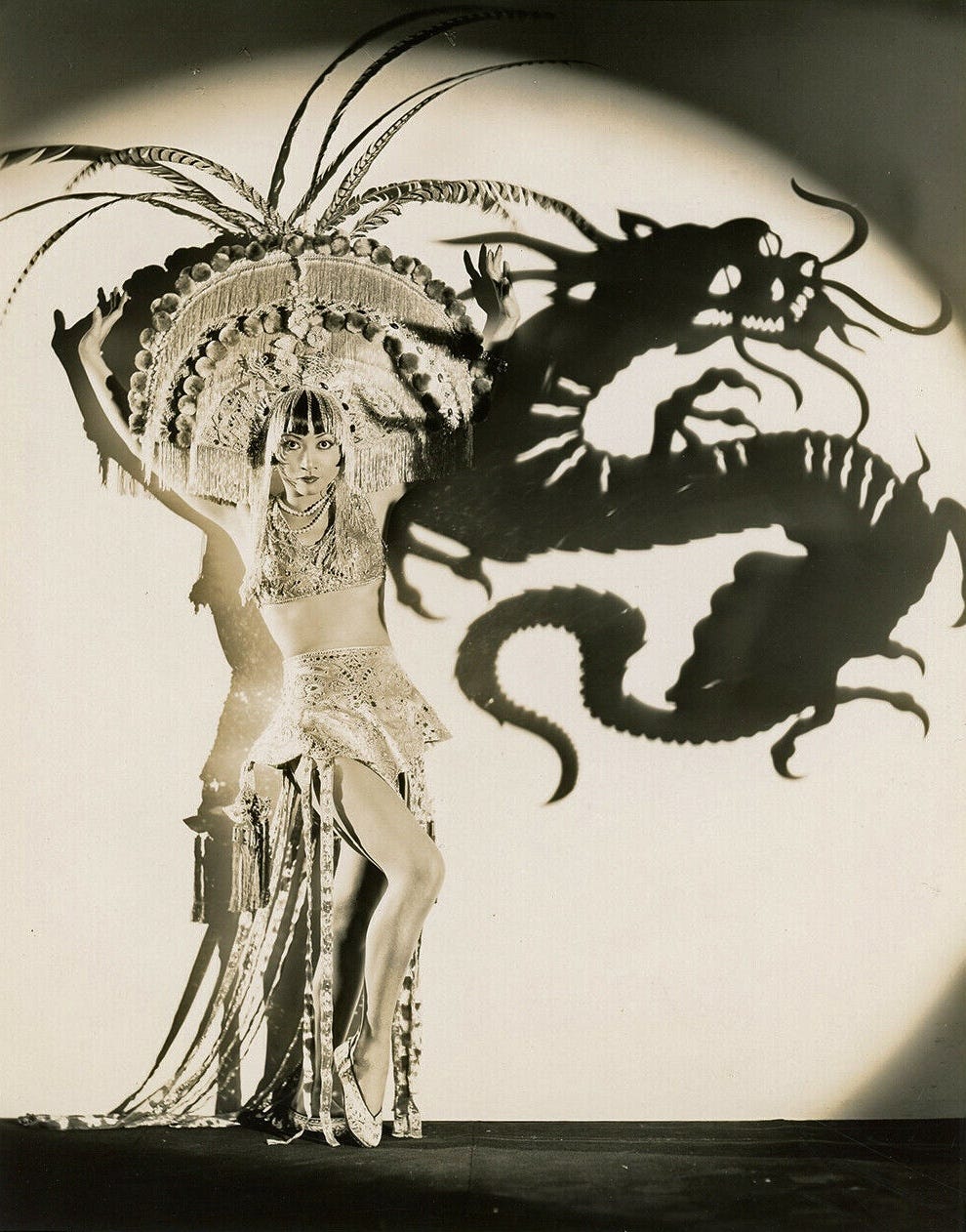 Anna May Wong poses against a wall in a shimmering showgirl outfit with a magnificent fringe headdress, a shadow of a Chinese dragon is projected onto the wall next to her