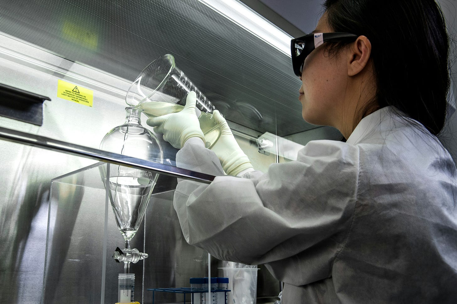 Woman wearing lab gear pouring liquid from one glass container to another behind a glass barrier