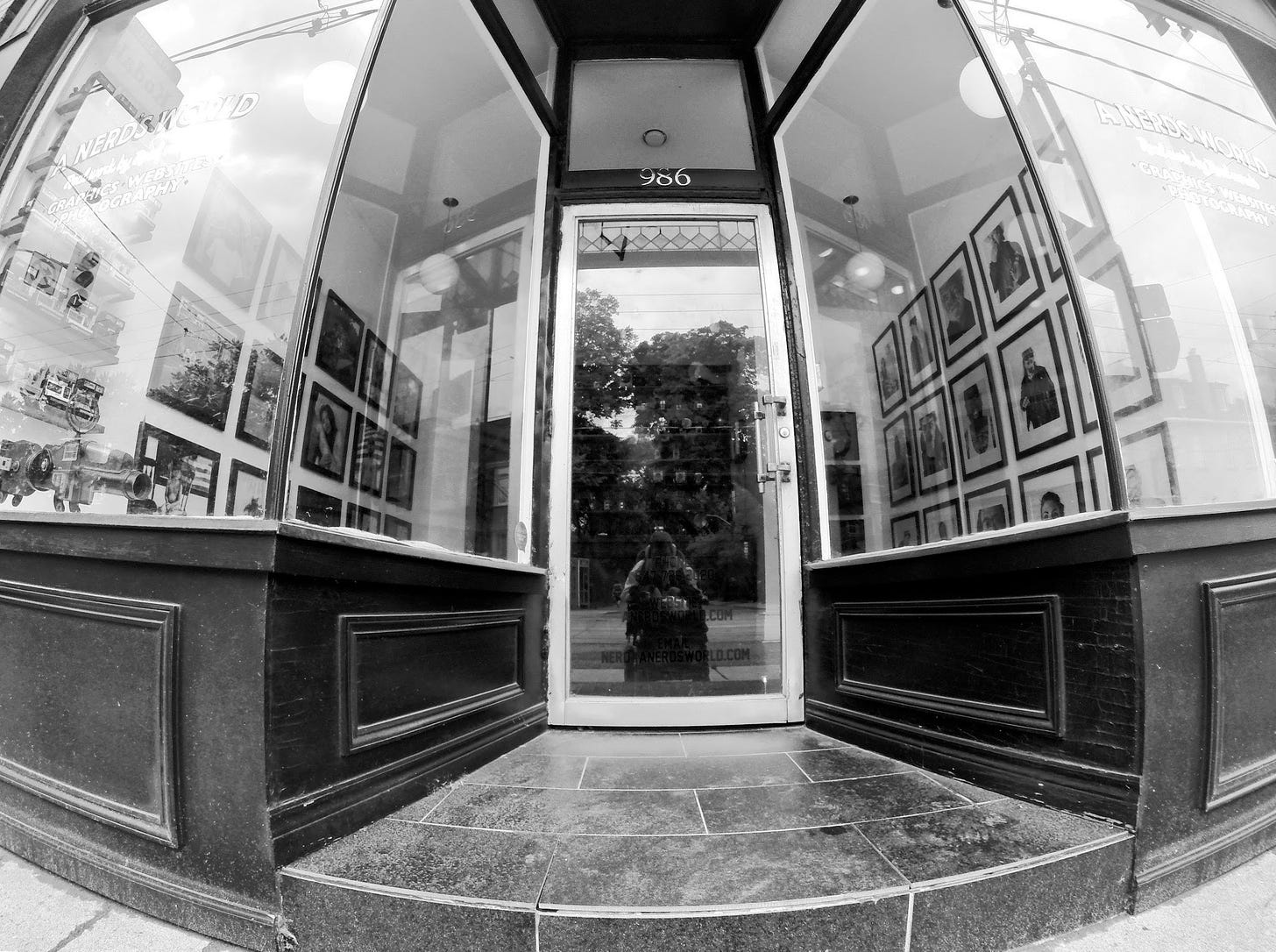 a symmetrical black and white image of Adam Cohoon sitting outside of a photography gallery with his reflection in the door. There is one step getting up into the gallery and beautiful photography hanging on the walls through the windows. Adam is sitting in his wheelchair with a GoPro camera, taking a long selfie to get into the gallery. 