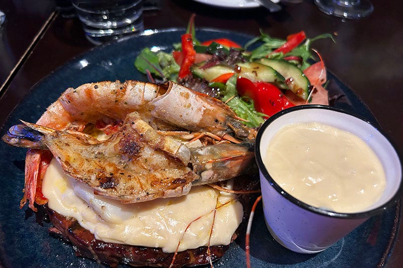 A steak on a plate with lots of cheese sauce, salad and prawns on top.