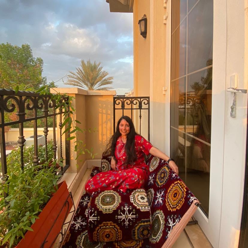 A picture of the writer in 2021 sitting outdoors in her balcony in the late afternoon sun with some fluffy clouds hovering above. Her wavy hair is down and she is wearing a red traditional clothes known as a "jalabiyya" in Arabic and is sitting on a modern version of a foldable Middle Eastern couch. She is nearly surrounded by plants.