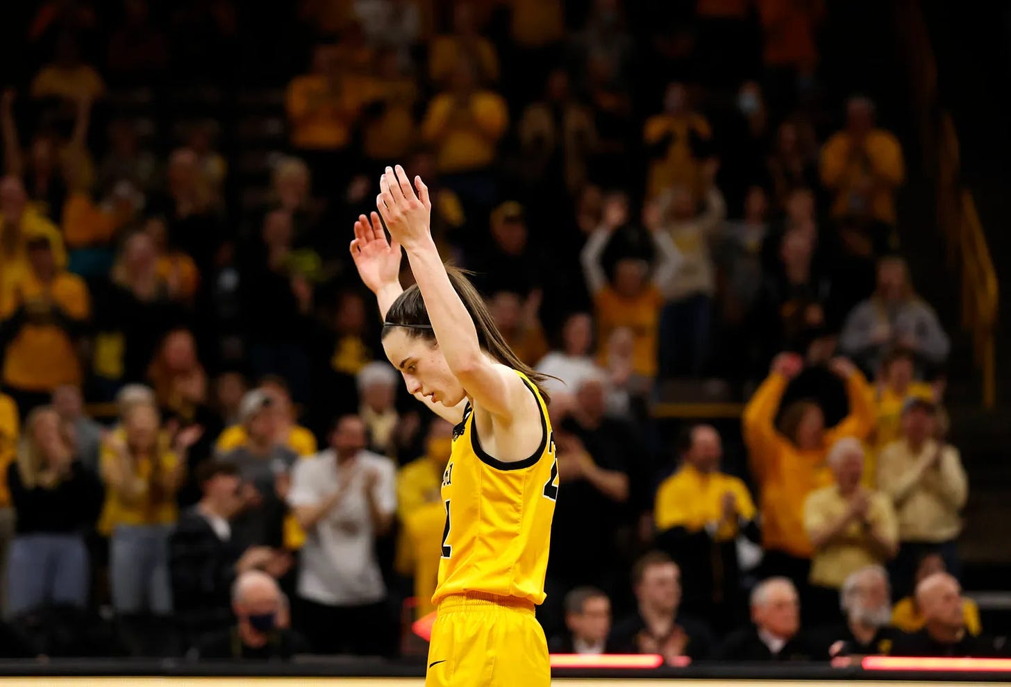 Caitlin Clark in a yellow Iowa jersey raises her hands to the sky after scoring.