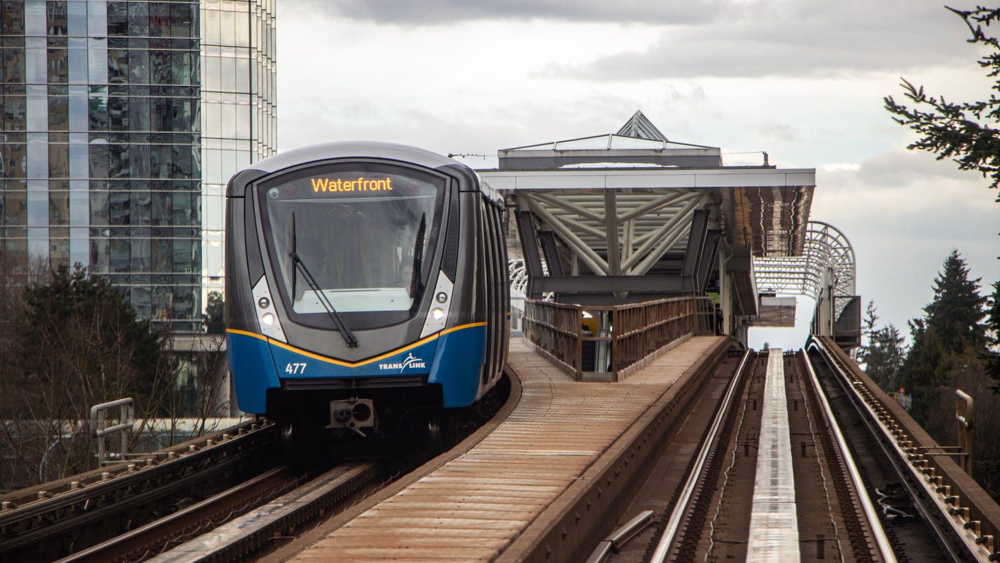 From the front of a SkyTrain approaching a train station. On the other side of the tracks, a train headed to Waterfront is leaving the station.