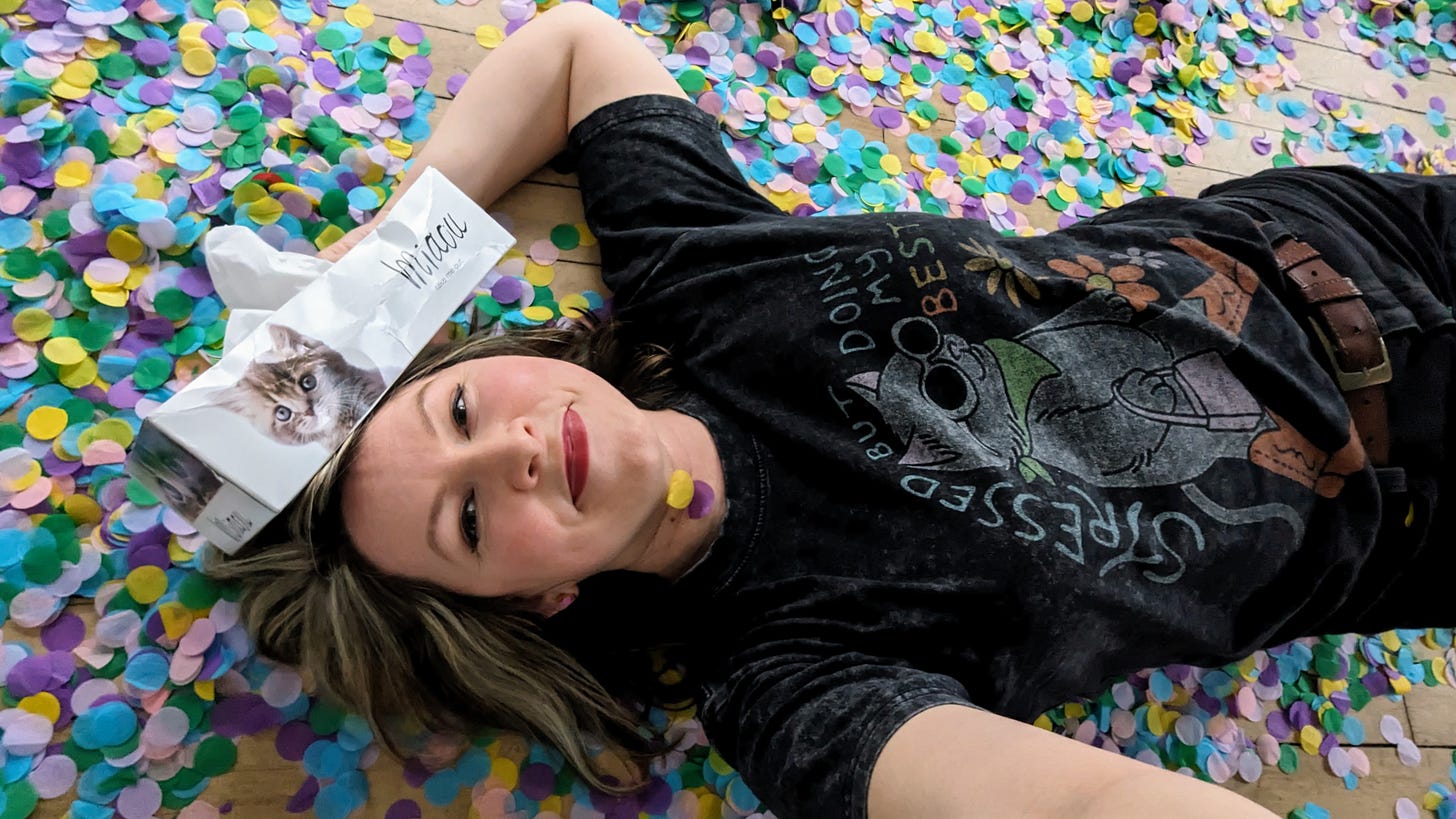 woman wearing cat t-shirt and holding tissue box with a cat on it, laying on the floor covered in colorful confetti
