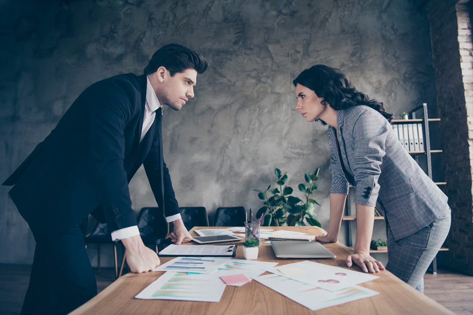 The image shows two professionals, a man and a woman, engaged in a serious discussion over documents on a table between them. Their body language and expressions indicate a potentially tense or critical interaction in a formal office setting.