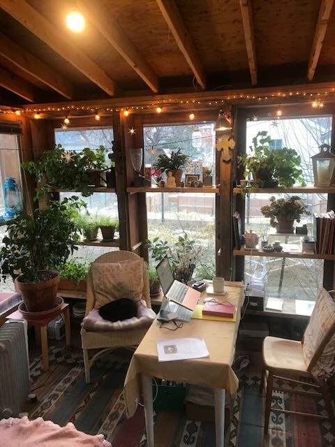 Shed roofed room with glass walls, shelves with plants, a chair with a sleeping cat, a small desk with manuscript, and a computer. 
