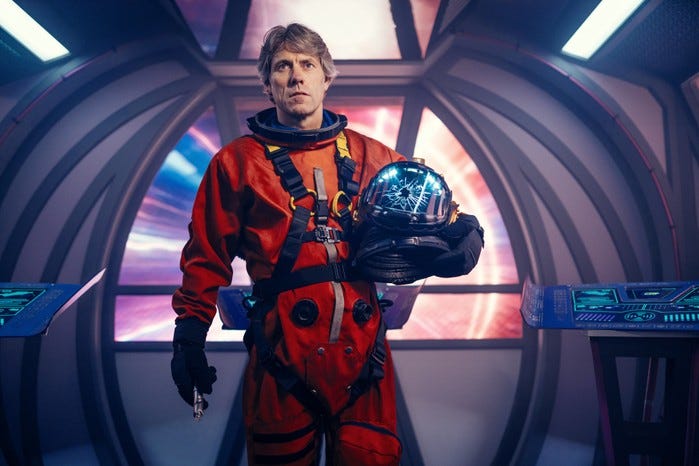 This image shows Dan Lewis (played by John Bishop) standing inside a bullet train racing through deep space, wearing an orange spacesuit, holding a helmet whose facemask has a hole blasted through it, and holding the Thirteenth Doctor's sonic screwdriver in Doctor Who Series 13's ninth episode, "The Power of the Doctor." This image shows Dan Lewis (played by John Bishop) standing inside a bullet train racing through deep space, wearing an orange spacesuit, holding a helmet whose facemask has a hole blasted through it, and holding the Thirteenth Doctor's sonic screwdriver in Doctor Who Series 13's ninth episode, "The Power of the Doctor."