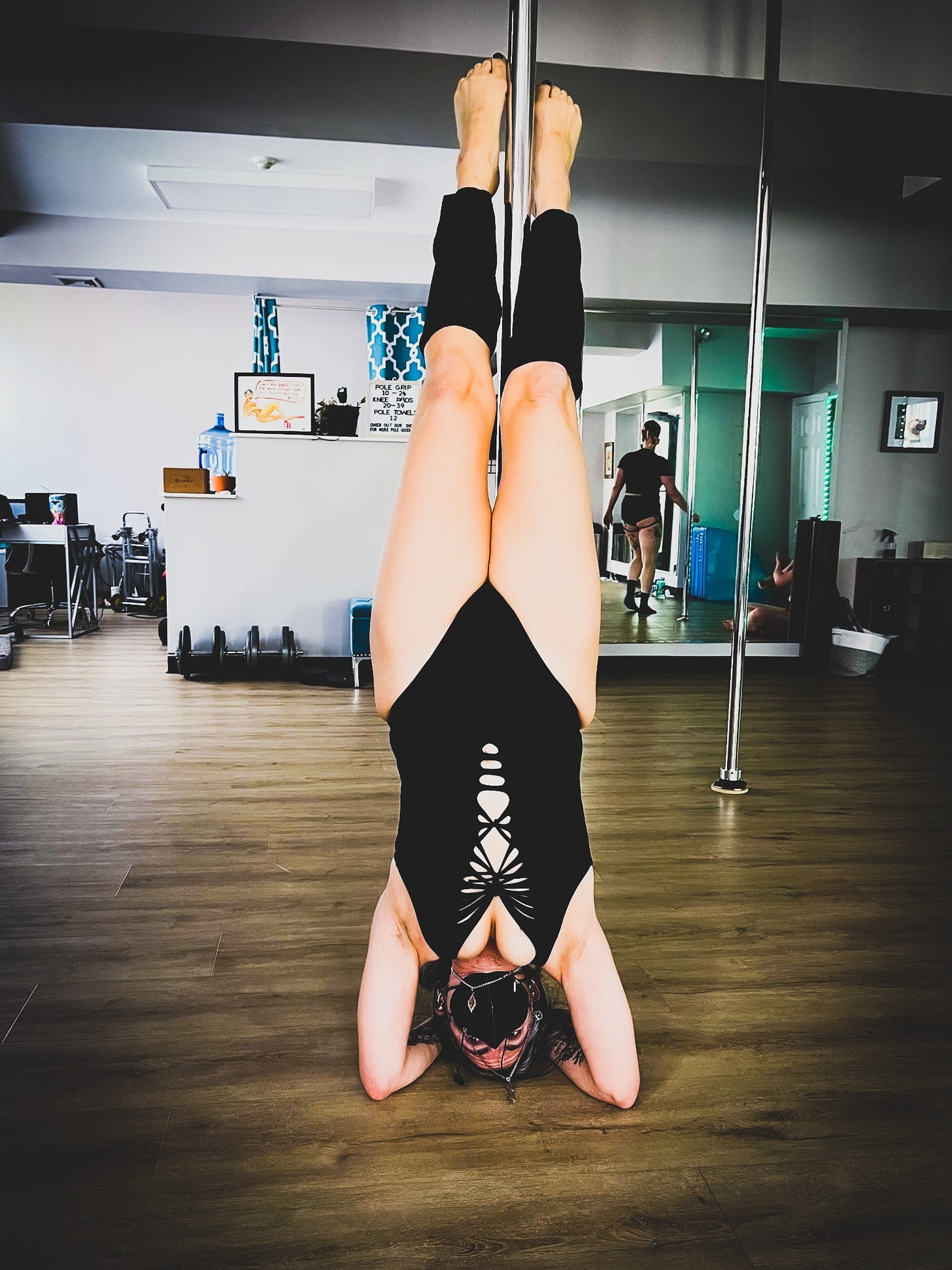 Woman doing headstand with a pole wearing a black leotard black leg warmers and a black face mask