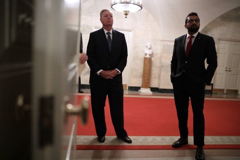 Two people are seen through an open door standing in a hallway.