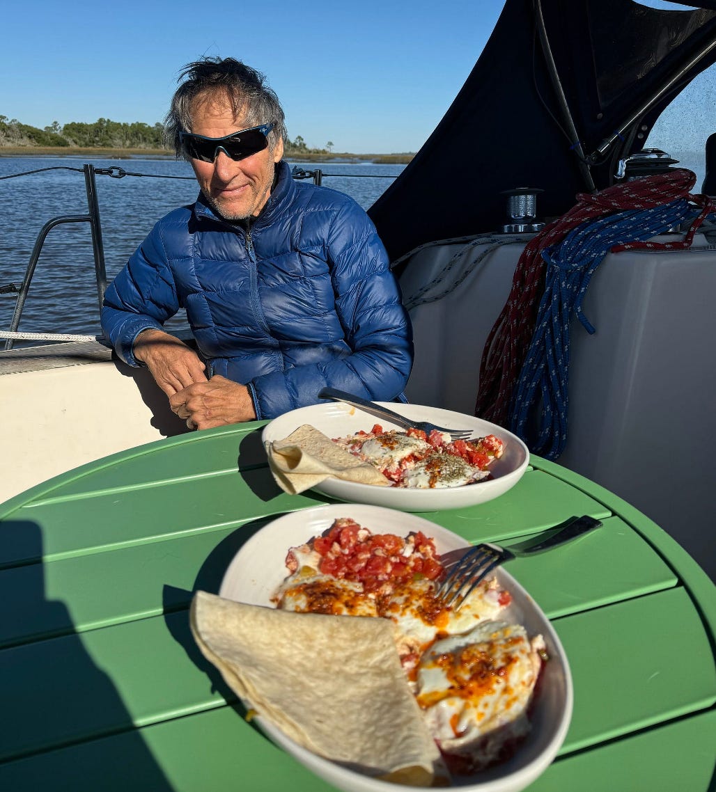 man sits with breakfast on a green table. Outside, blue water, in a boat.