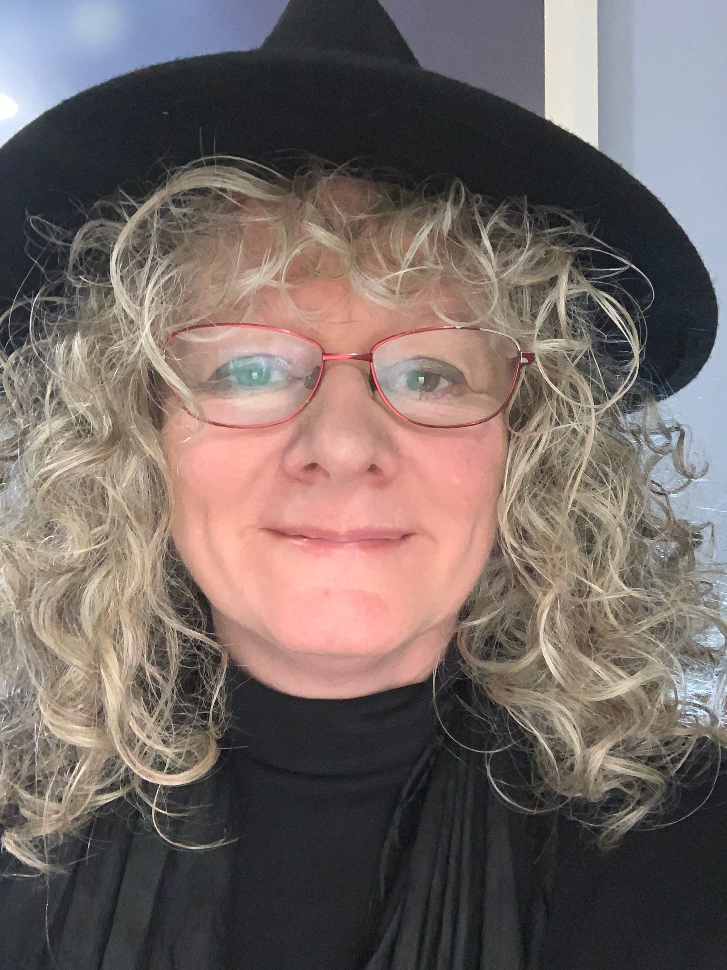 A woman’s face, with silver curly hair, red glasses and a black hat. 