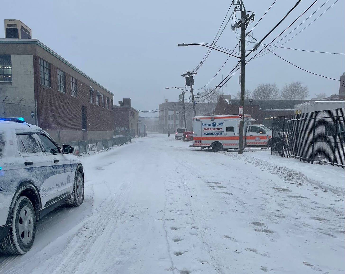A view of Atkinson street blanketed with an inch of snow as more snowflakes come down. The street is clear except for a police cruiser, ambulance, and staff member walking at the end of the street