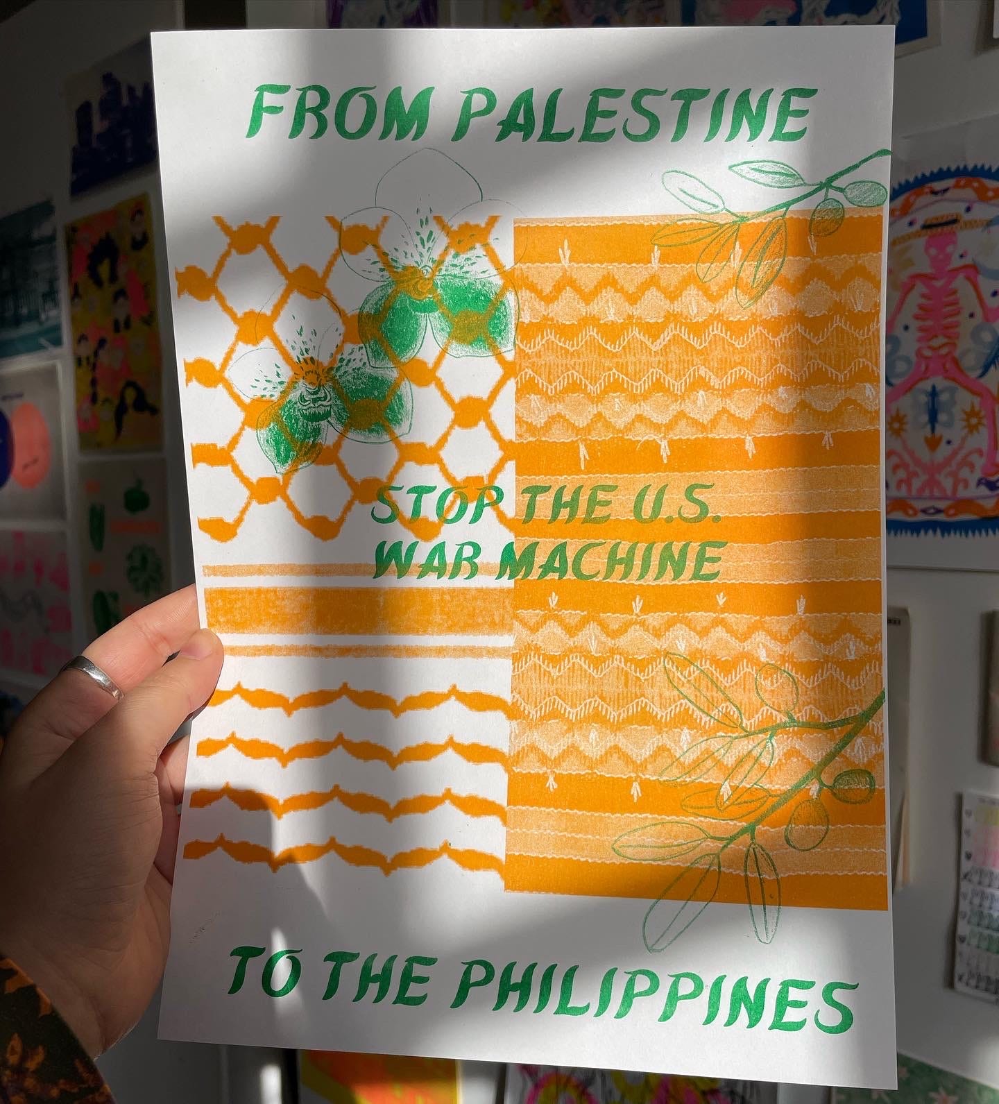 photo of me holding a green and orange poster that reads "FROM PALESTINE TO THE PHILIPPINES, STOP THE US WAR MACHINE" in green Filipino style calligraphy. There is a keffiyeh and Filipino indigenous fabric print side by side in orange. Drawings of two orchids and two olive branches are overlayed in green.