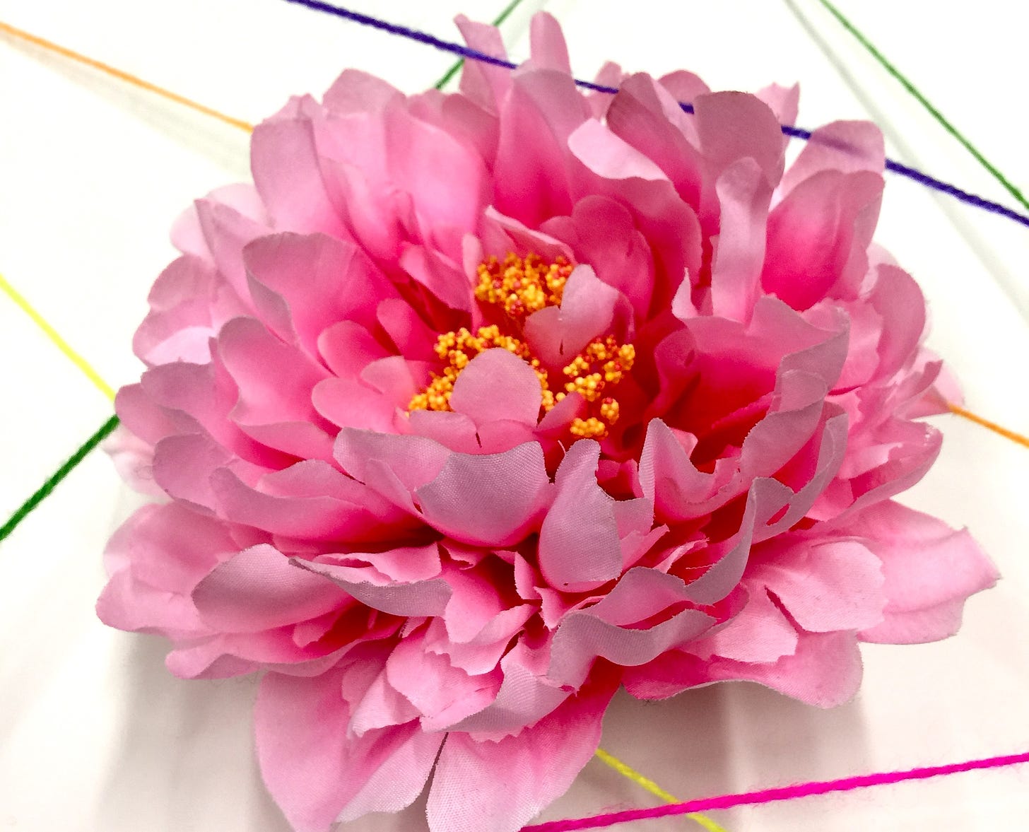 A bright pink artificial peony sits against a white background with crossing lines of colored yarn.
