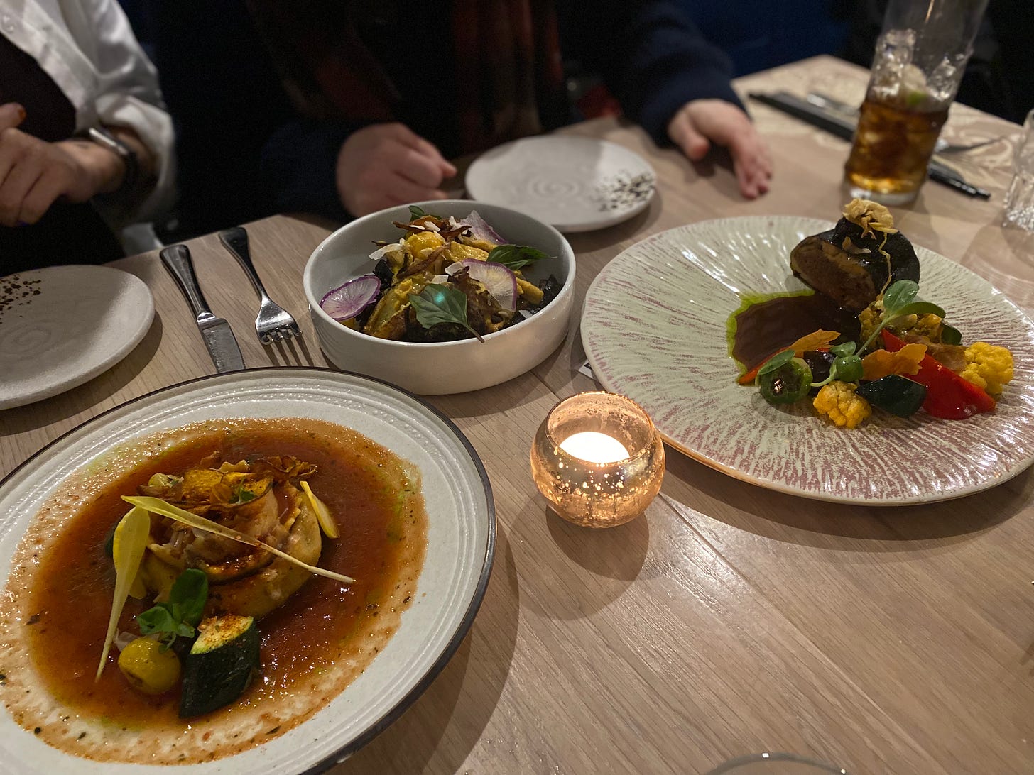 Three dishes of different sizes and shapes on a table with a votive candle in the centre. One features a large stuffed pasta in a thin sauce, one holds a vegetable curry over black rice, and the last has a mushroom roulade with roasted vegetables, a jus in the bottom of the plate. Two people's hands are visible in the background, in front of side plates and cutlery.