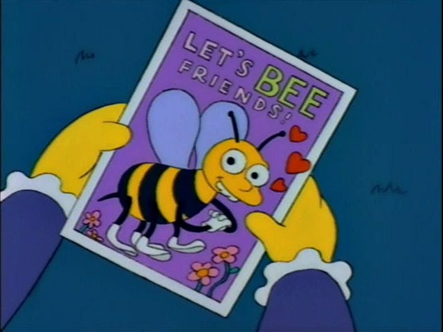 Ralph Wiggum from The Simpsons holding a Valentine’s Day card that says, “Let’s BEE friends!” The word “bee” is spelled like honeybee.