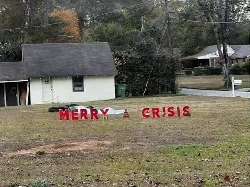 White home with gray roof, in front is a green grass yard, with sign that says "Merry Crisis" in red letters