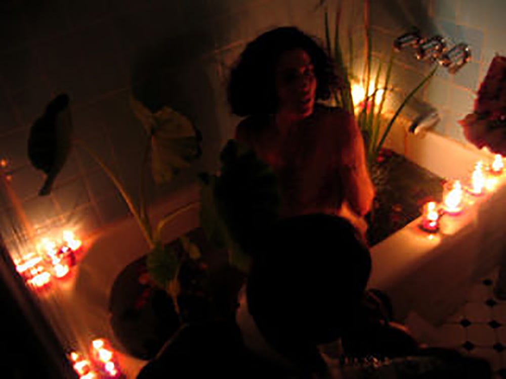 A darkened, slightly blurred image of a bathtub with plants and flowers in the water and red candles around the rim. The Love Artist is in the bath, and you can just make out the sillhouette of another person kneeling next to the tub. 