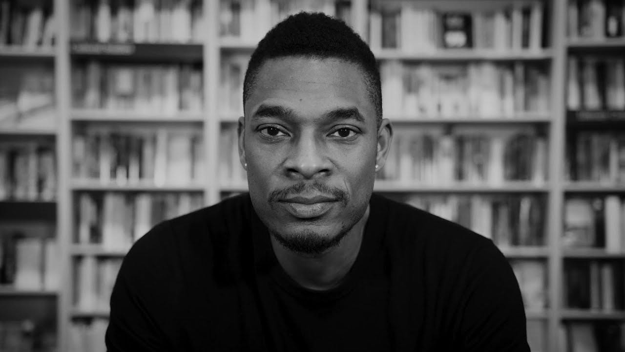 A black and white photo of Terrance Hayes. He looks directly into camera, smiling very slightly. Behind him, out of focus, are shelves filled with books.