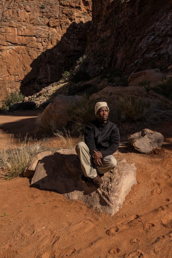 A man wearing a black jacket, tan pants and a tan hat sits atop a large boulder that’s partially embedded in a red sandy soil. Behind him, dramatically red-brown cliff faces rise from the earth.