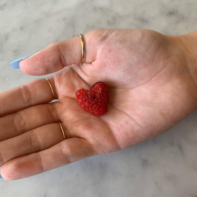 raspberry in the shape of a heart had in a palm with long blue acrylic nails and gold rings on the fingers above a white stone surface