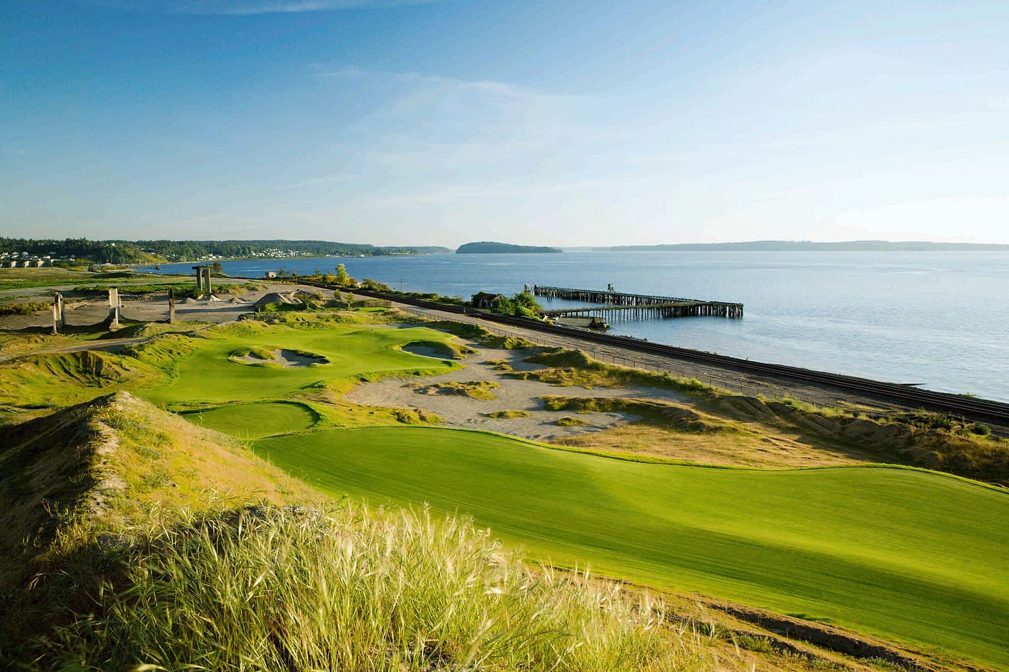 Aerial view of Chambers Bay Golf Course showing undulating fairways and sandy waste areas along Puget Sound. The course's natural contours and fescue rough create a striking contrast with the calm waters of the sound stretching to distant mountains. Aerial view of Chambers Bay Golf Course showing undulating fairways and sandy waste areas along Puget Sound. The course's natural contours and fescue rough create a striking contrast with the calm waters of the sound stretching to distant mountains.