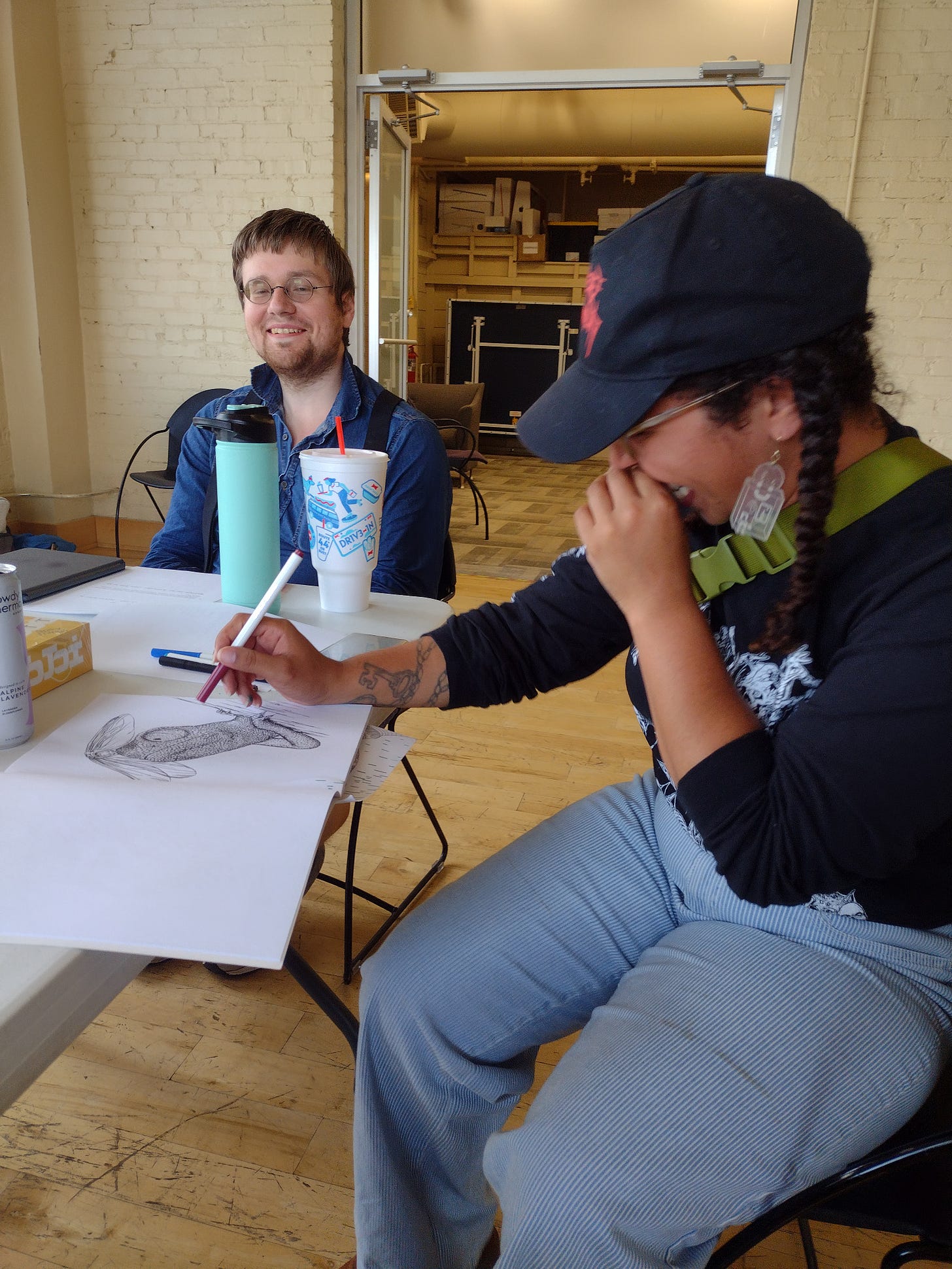 Two people are sitting in chairs at a table. One person is smiling and coloring in a coloring book, and the other person is smiling and looking straight into the camera.