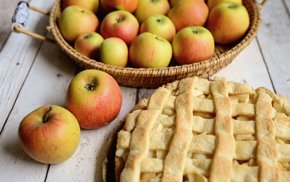 Photo of a basket of apples and 2 loose apples on a wooden table with a baked, lattice-topped apple pie