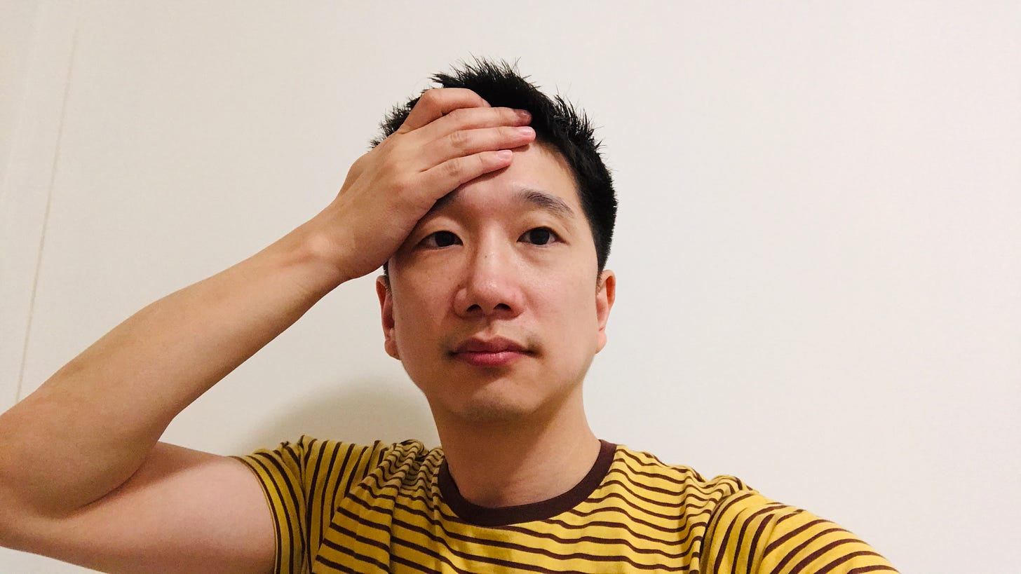 Anton selfie against a white wall wearing a yellow shirt with brown stripes. He has a hand to his head.