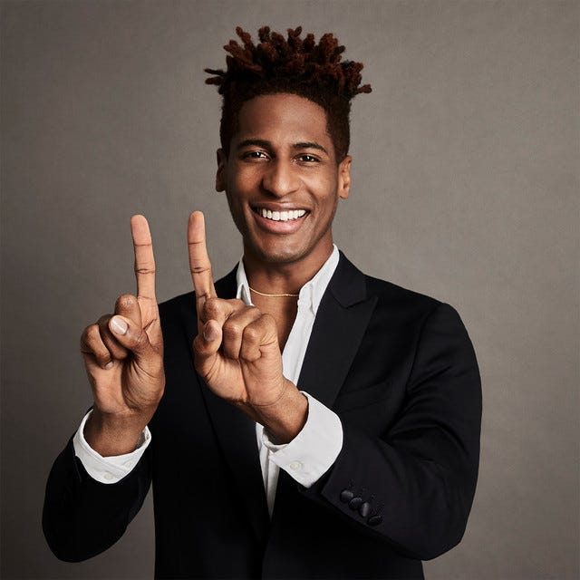 Jon Batiste smiling and holding up one finger each from both hands