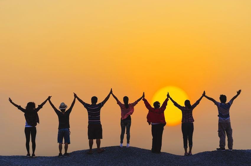 Seven people holding hands while looking at the sunset. Their hands are held above their heads, looking like the letter Y.