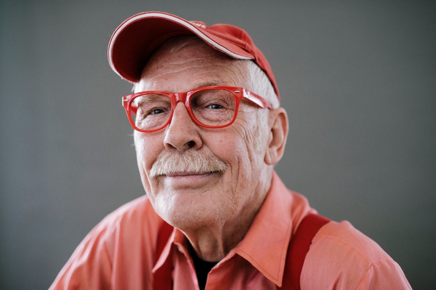 Older, smiling white man with white mustache looks into camera in this portrait.. He wears a red cap, red glasses and red shirt.