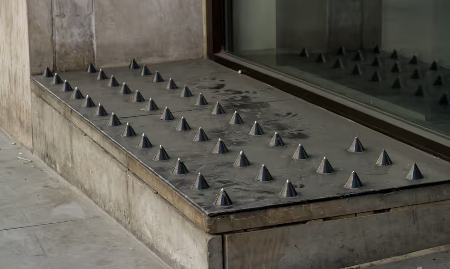 Photo of a level area on the street in front of a building that has many menacing spikes added to the surface to keep people from being able to sit or sleep on it.