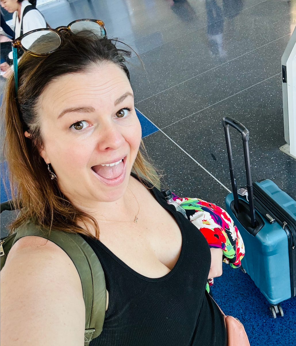 A selfie of Amber at the airport. She wears a backpack and there is a small, blue suitcase behind her.