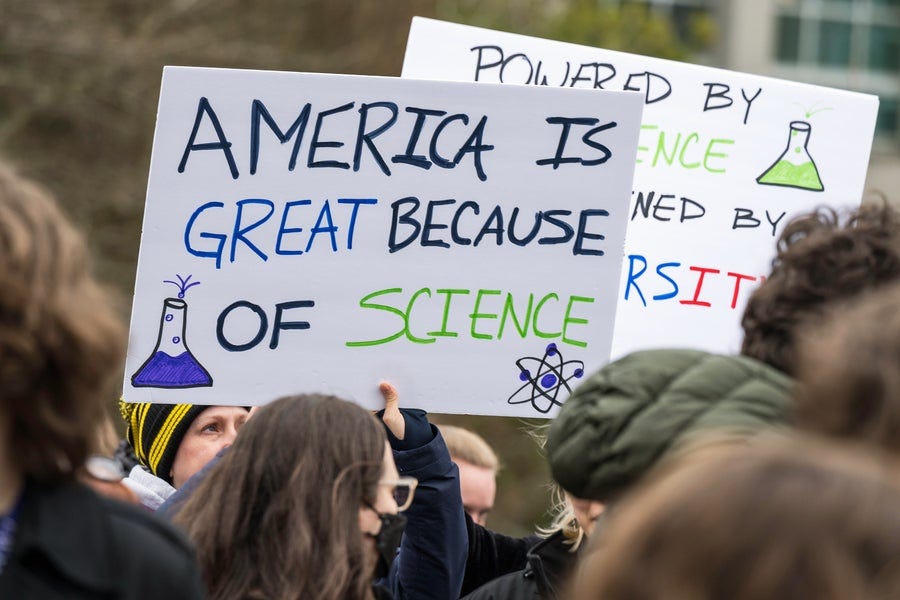 Participants attending the Hands Off Our Healthcare, Research and Jobs rally at University of Washington Portage Bay Campus in Seattle hold signs. In the foreground a sign reads, "America is great because of science." Participants attending the Hands Off Our Healthcare, Research and Jobs rally at University of Washington Portage Bay Campus in Seattle hold signs. In the foreground a sign reads, "America is great because of science."