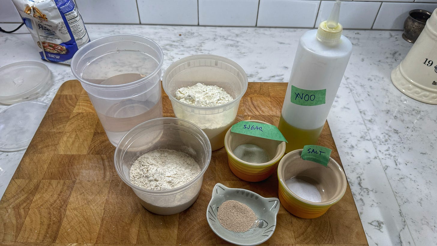 A collection of ingredients for pizza crust on a cutting board. Clockwise from top left, they incude: water, flour, sugar, olive oil, salt, yeast, and crokkia flour