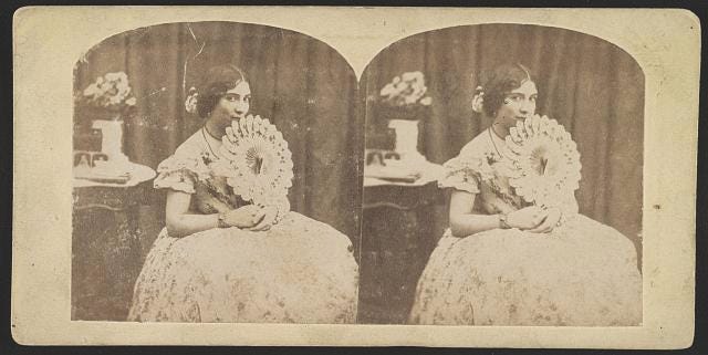 Photograph shows a woman in a formal dress sitting, holding a fan.