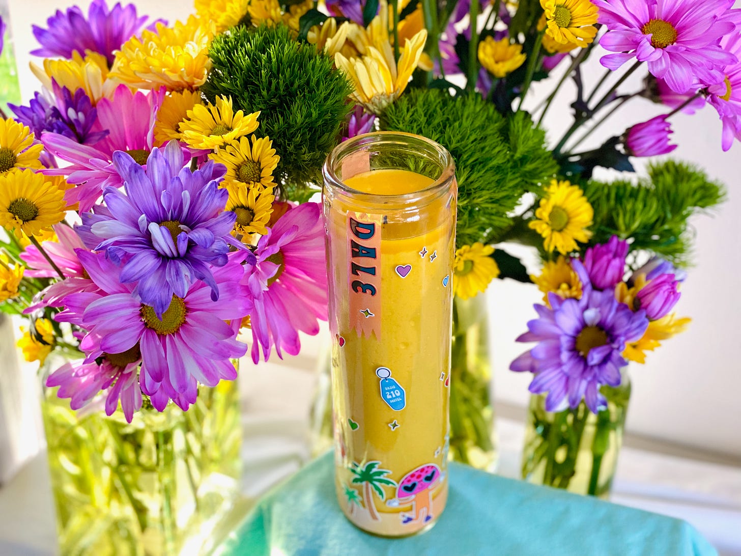 photo of a gaudy prayer candle covered with stickers nestled among a riot of tacky, bright flowers.