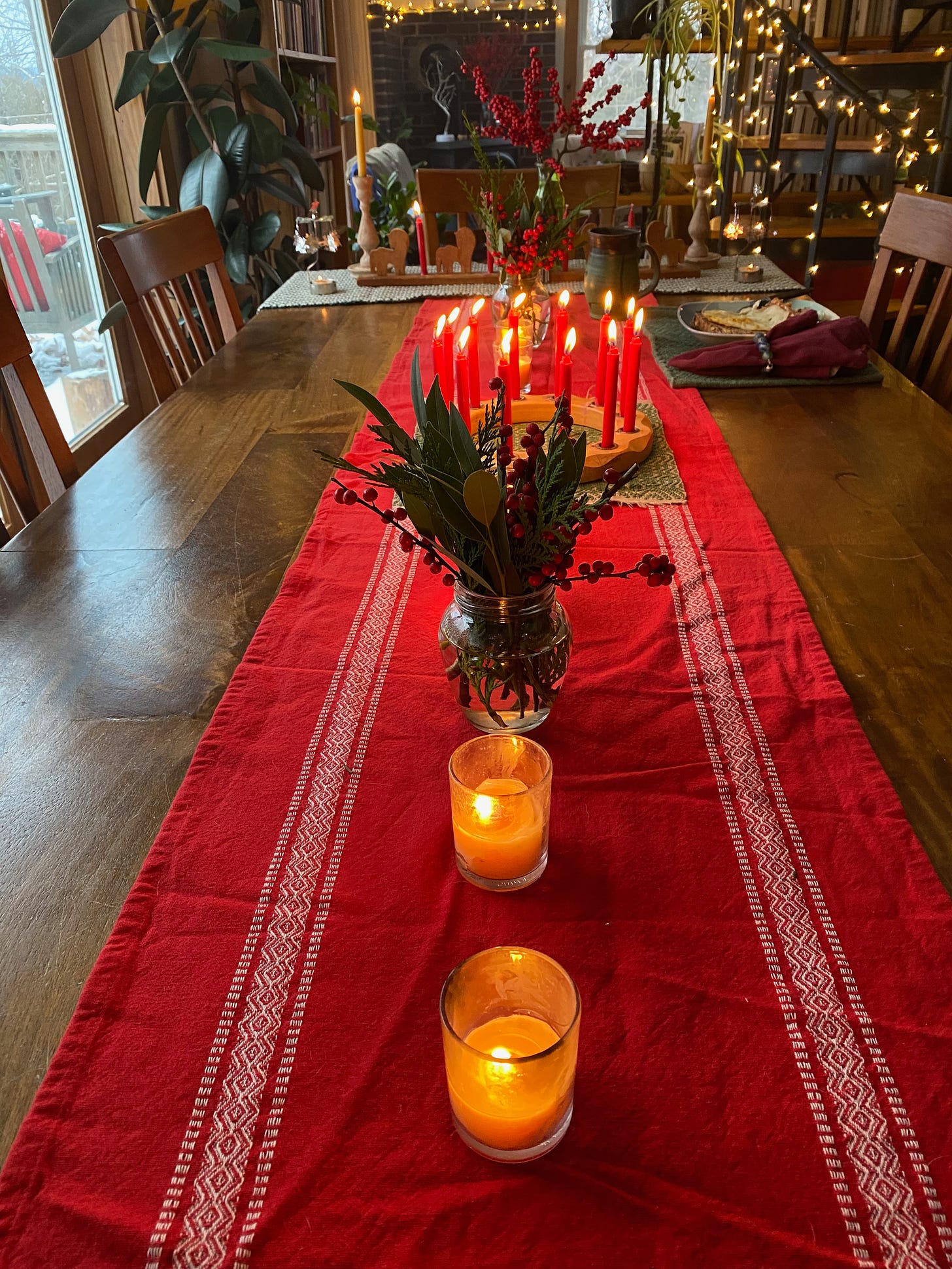 A table decorated with a red runner, jars of winterberry and greenery, and many lit candles.